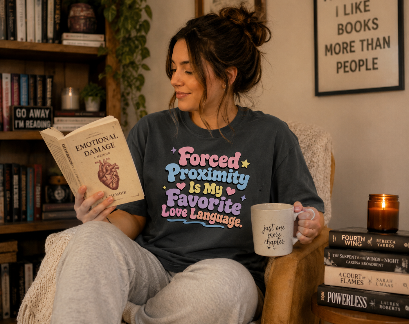 Woman sitting in a cozy room holding a book and mug, wearing a shirt with a colorful text design.